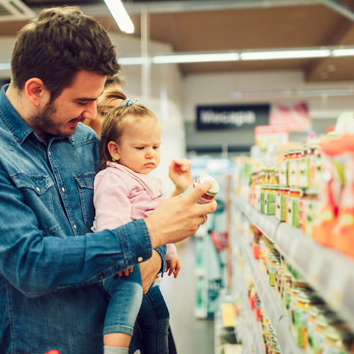 Father with toddler at a supermarket shelf. The toddler looks discontentedly at a glass package.